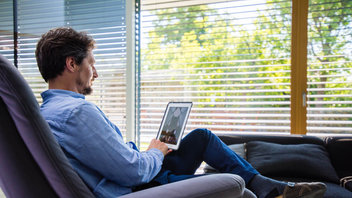 Mann sitzt im Wohnzimmer und steuert die Fensterrolläden per Smart-Home-Steuerung am Tablet