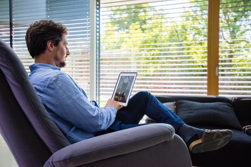 Mann sitzt im Wohnzimmer und steuert die Fensterrolläden per Smart-Home-Steuerung am Tablet